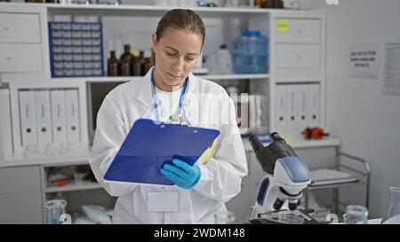 Jeune scientifique blonde focalisée, presse-papiers à la main, mène diligemment des recherches médicales précises dans un laboratoire animé, se plongeant dans le cru Banque D'Images