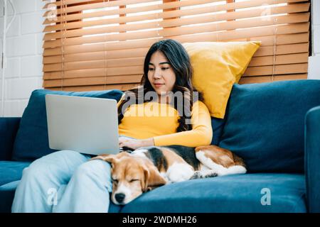 Une jolie jeune femme, ordinateur portable à la main, est assise sur son canapé à la maison. Son chien Beagle dort paisiblement Banque D'Images