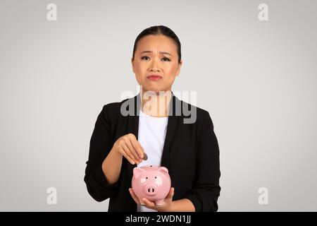 Inquiète femme d'affaires asiatique dans un blazer noir dépose une pièce dans une tirelire Banque D'Images