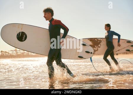 Groupe de surfeurs heureux en combinaison courant dans la mer tenant des planches de surf Banque D'Images