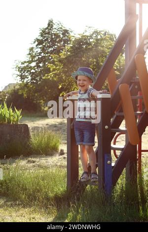 Heureux petit garçon posant et souriant sur les marches en bois de la maison de jeu. Une belle et merveilleuse soirée d'été chaude. Lumière du soir, photo verticale. Banque D'Images