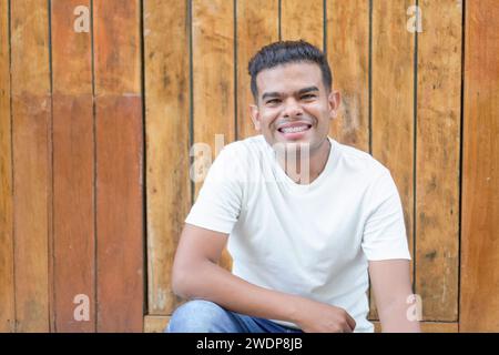 Jeune homme avec des appareils dentaires souriant et assis avec désinvolture près d'un mur de lattes en bois. Banque D'Images