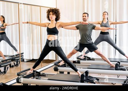 Groupe de jeunes athlètes masculins et féminins en vêtements actifs faisant pilates pose debout avec les bras tendus sur des lits de reformeur dans la salle de gym Banque D'Images