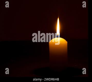 La flamme ou la lumière de bougie brûlante unique brille sur une grande bougie blanche sur fond noir ou sombre sur la table dans l'église pour Noël, funérailles ou mémorial Banque D'Images