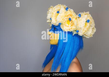 Vue de côté Portrait en gros plan créatif d'une jeune femme avec un art du visage jaune et bleu, une couronne de pivoines sur sa tête sur un fond gris isolé. C Banque D'Images