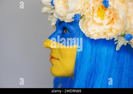 Vue de côté Portrait en gros plan créatif d'une jeune femme avec un art du visage jaune et bleu, une couronne de pivoines sur sa tête sur un fond gris isolé. C Banque D'Images