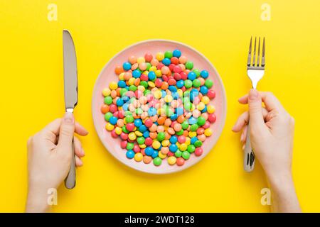 la fille tient des couverts dans ses mains et mange des bonbons dans une assiette. Concept de santé et d'obésité, vue de dessus sur fond coloré. Banque D'Images