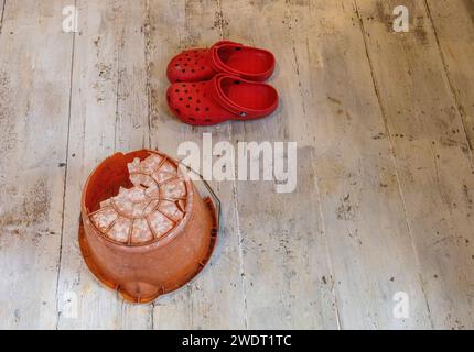 Sur des planches blanchies à la chaux, un seau en plastique orange inversé avec un énorme trou dans le fond et une paire de Crocs rouge vif Banque D'Images