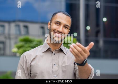 Joyeux jeune homme afro-américain dans une tenue d'affaires décontractée debout en toute confiance devant un immeuble de bureaux moderne. Banque D'Images