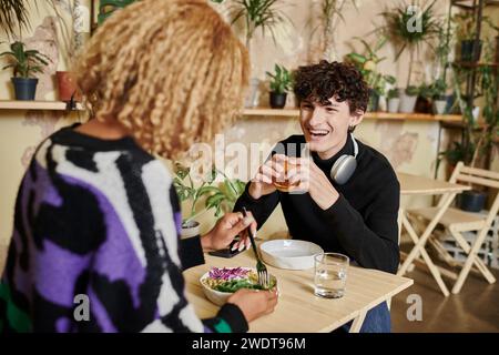 heureux homme bouclé mangeant un hamburger de tofu près d'une petite amie afro-américaine appréciant une salade dans un café végétalien Banque D'Images