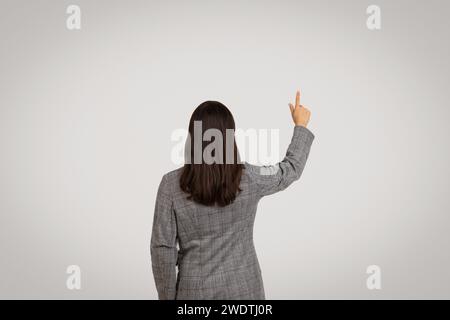 Vue arrière de femme d'affaires pointant sur l'écran virtuel, touchant le bouton invisible, fond de studio gris Banque D'Images