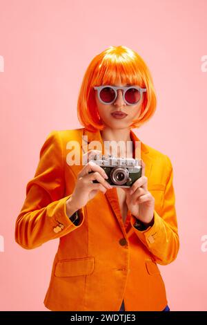 Mode dynamique. Jeune femme dans les lunettes de soleil à la mode habillé blazer orange, avec perruque regardant la caméra et tient la caméra photo contre rose pastel Banque D'Images