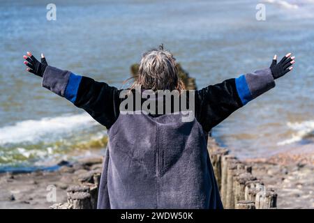 Femme mûre se tient avec les bras écartés, regardant la mer par derrière, bravant la journée froide avec sérénité contemplative. Banque D'Images