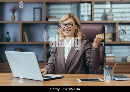 Joyeuse femme d'affaires mature avec des lunettes travaillant sur ordinateur portable dans un bureau à domicile moderne, exprimant la confiance et le professionnalisme. Banque D'Images
