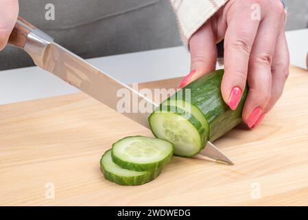 Trancher le concombre en tranches sur une planche à découper en bois. Les mains féminines coupent un concombre avec un couteau. Cuisine maison. Banque D'Images