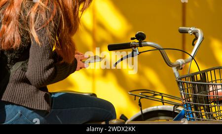Dans les rues de la ville, une japonaise, près d'un vélo, engage une conversation textuelle sur son smartphone avec un fond jaune. Une dame asiatique o Banque D'Images