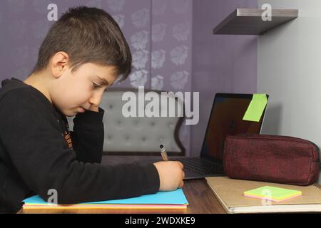 Enseignement à distance en ligne. Un écolier étudie à la maison et fait ses devoirs scolaires. Étudiant ennuyé essayant d'écrire de la composition. Reste à la maison. Banque D'Images