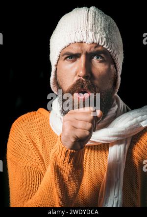 Homme malade barbu en chandail, chapeau et écharpe toussant. Beau gars ayant mal à la gorge et toussant dans son poing. Homme malsain enveloppé dans une écharpe ont Banque D'Images