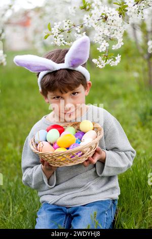 Chasse aux œufs de Pâques. Mignon garçon préscolaire portant des oreilles de lapin tenant le panier plein d'oeufs de Pâques dans les mains à la chasse aux oeufs de Pâques dans le jardin. Portrait contre le dos Banque D'Images