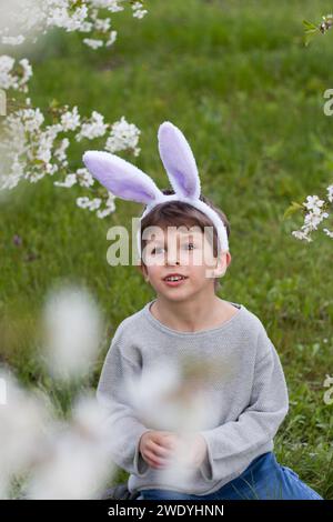 Mignon garçon préscolaire avec des oreilles de lapin dans le jardin. enfant souriant assis sur la pelouse verte sur fond d'arbres fleurissant avec des fleurs blanches. Joyeuses pâques Banque D'Images