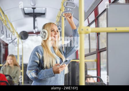 Vue de côté de jolie fille blonde debout avec mobile regardant la fenêtre souriante dans les transports en commun. Récolte de poignée de maintien femelle avec des passagers brouillés assis dans le bus. Concept de voyage. Banque D'Images