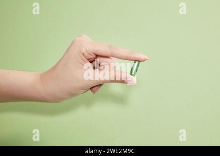 Une main féminine tenant une seule capsule verte de vitamine sur un fond vert clair. Concept de médicament avec espace à copier. Banque D'Images