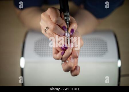 Gros plan détaillé d'une femme méconnaissable enlevant son propre vernis à ongles avec une lime électrique sur un dépoussiéreur, bricolage manucure gros plan Banque D'Images