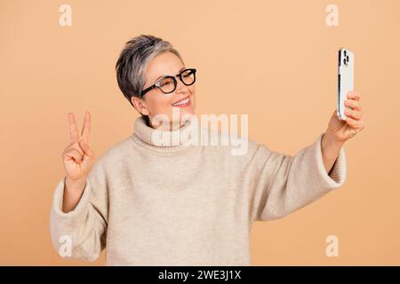 Photo de femme d'affaires influenceuse retraitée dame fait signe v créer un contenu d'autoportrait pour instagram isolé sur fond de couleur beige Banque D'Images