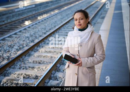 Jeune femme d'affaires adulte tenant le téléphone portable en attendant le train aérien à la station de métro dans une ville, debout à la station de quai de transpo public Banque D'Images