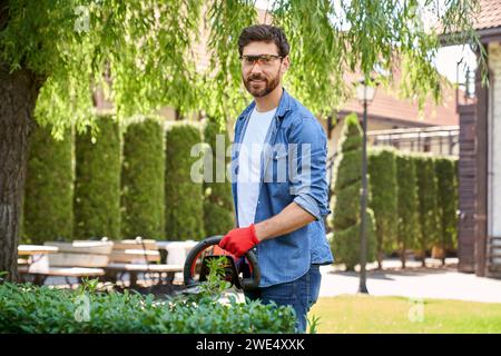 Bel homme portant des lunettes de sécurité élaguant buisson avec cutter dans le parc. Portrait d'un jardinier expérimenté coupant le dessus des haies avec des coupe-branches électriques et regardant la caméra. Concept de lieu de travail. Banque D'Images