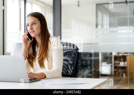 Une femme d'affaires concentrée converse sur son téléphone portable tout en utilisant un ordinateur portable, au cours d'une discussion sur des questions commerciales importantes avec le fond de bureau moderne avec de grandes fenêtres Banque D'Images