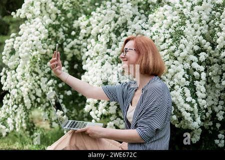 Style de vie freelance, travail à distance en pleine floraison une jeune femme capture son moment parmi les fleurs blanches avec un ordinateur portable. Banque D'Images