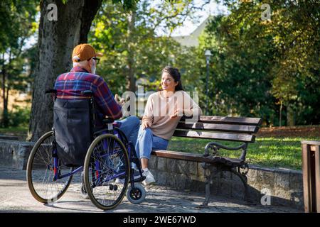 Soignant souriant assis sur un banc écoutant un homme retraité en fauteuil roulant au parc Banque D'Images