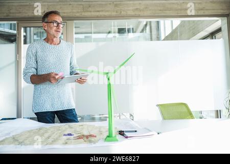 Homme d'affaires réfléchi debout avec tablette PC près du modèle d'éolienne au bureau Banque D'Images