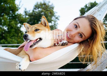 Femme blonde souriante couchée avec le chien Shiba Inu dans un hamac Banque D'Images