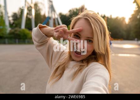 Femme heureuse prenant selfie et gestuant signe de paix dans le parc d'attractions au coucher du soleil Banque D'Images