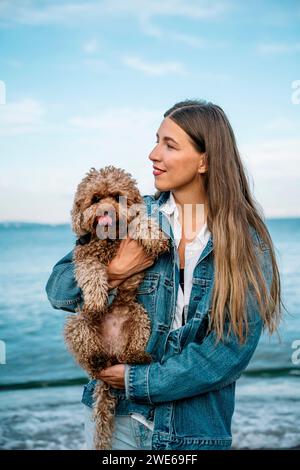 Jeune femme souriante avec chien caniche debout à la plage Banque D'Images
