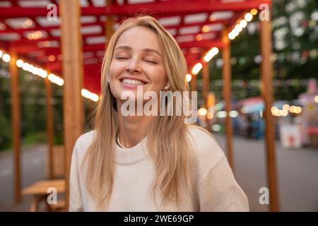 Jeune femme blonde souriante portant des écouteurs intra-auriculaires sans fil écoutant de la musique au parc d'attractions Banque D'Images