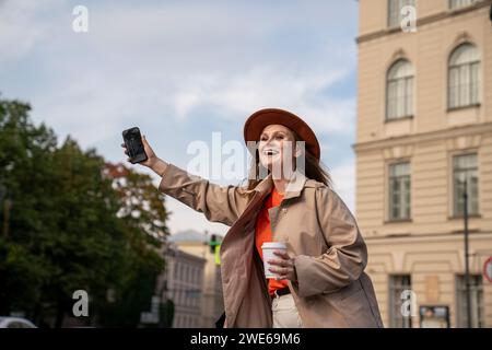 Femme heureuse tenant le téléphone intelligent et hailing tour près du bâtiment Banque D'Images