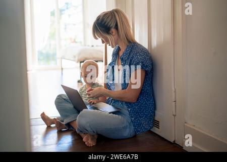 Mère qui travaille avec un ordinateur portable prenant soin de son fils assis à la maison Banque D'Images