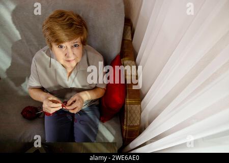 Femme senior tricotant sur une chaise près du rideau à la maison Banque D'Images