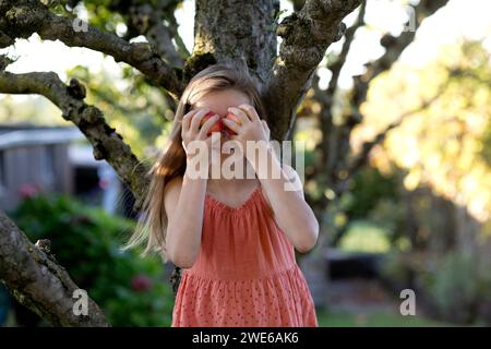 Fille souriante tenant des fruits de pomme sur le visage dans le jardin Banque D'Images