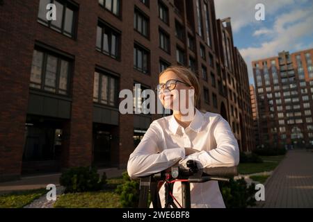 Femme d'affaires souriante avec scooter de poussée électrique par le bâtiment Banque D'Images