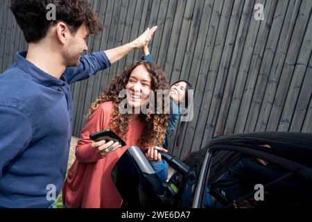 Homme et femme faisant high-five par un ami chargeant la voiture électrique Banque D'Images