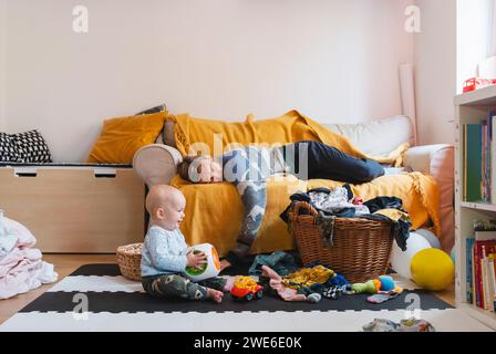 Mère épuisée dormant sur le canapé avec bébé garçon jouant à la maison Banque D'Images