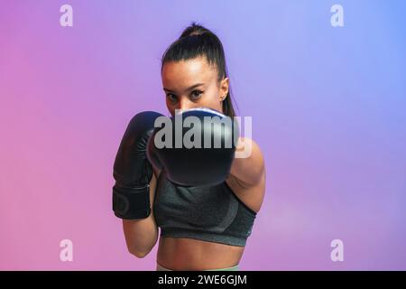 Jeune femme confiante avec des gants de boxe noirs s'exerçant sur fond coloré Banque D'Images