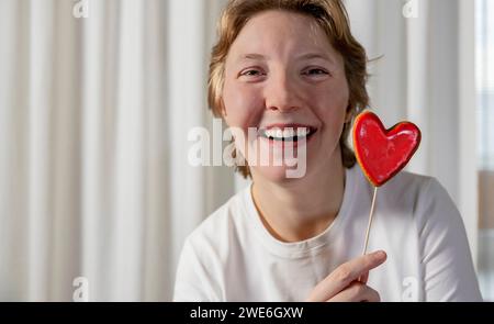 Heureuse jeune femme avec le biscuit en forme de coeur devant le rideau blanc Banque D'Images