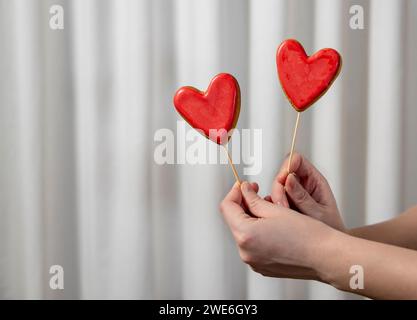 Mains de femme tenant des biscuits en forme de coeur rouge devant le rideau blanc Banque D'Images