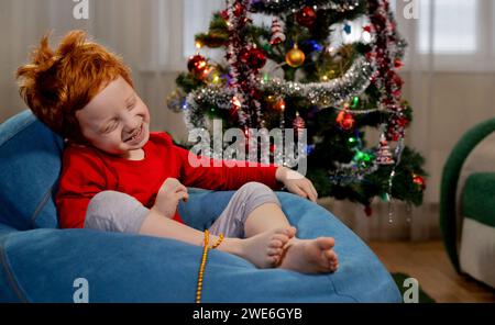 Joyeux garçon roux assis sur la chaise de sac de haricot bleu près de l'arbre de Noël à la maison Banque D'Images