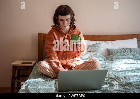 Fille regardant un film de série sur un ordinateur portable assis dans le lit boire du thé à la maison tranquillement passant le temps le jour de congé Banque D'Images